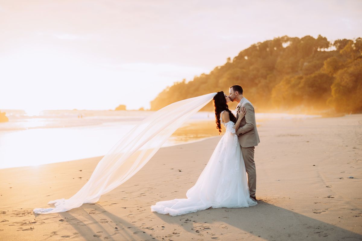 A couple in wedding attire embrace on a misty beach at sunset as the bride's veil flows in the wind.
