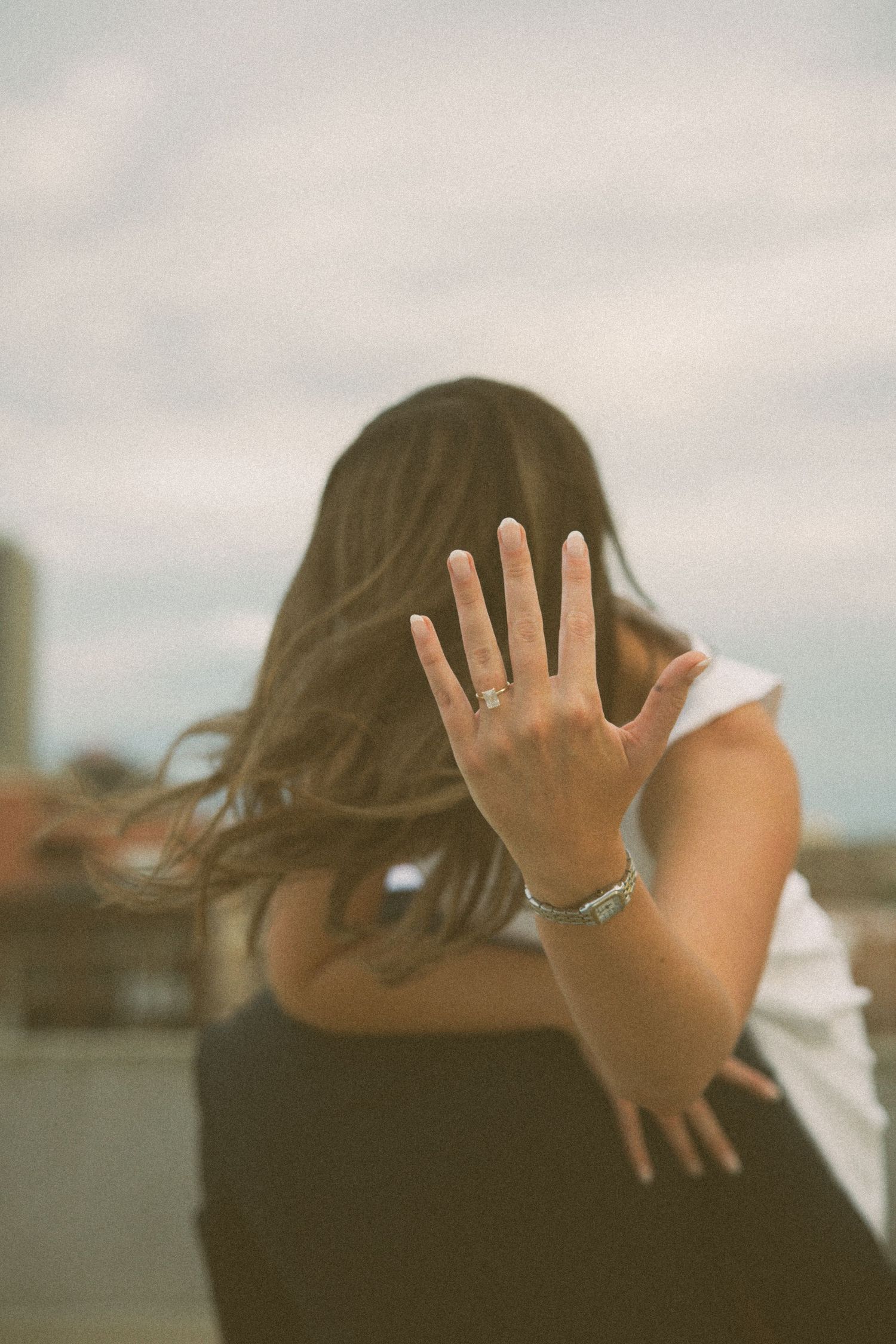 A person shows off their engagement ring while covering their face with long hair flowing.