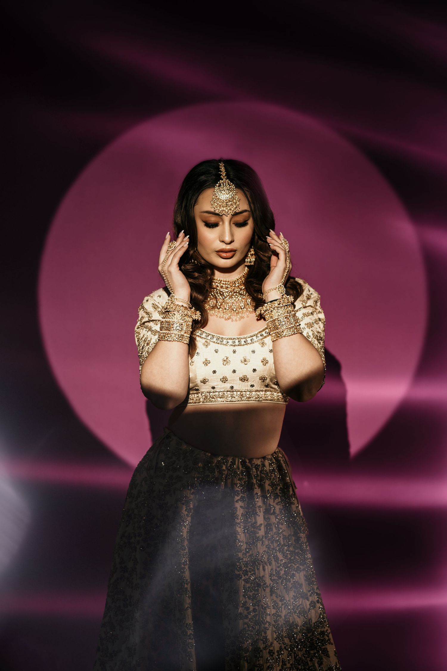 Traditional South Asian bride in ornate gold jewelry and lehenga poses against dramatic purple backdrop with moon silhouette.