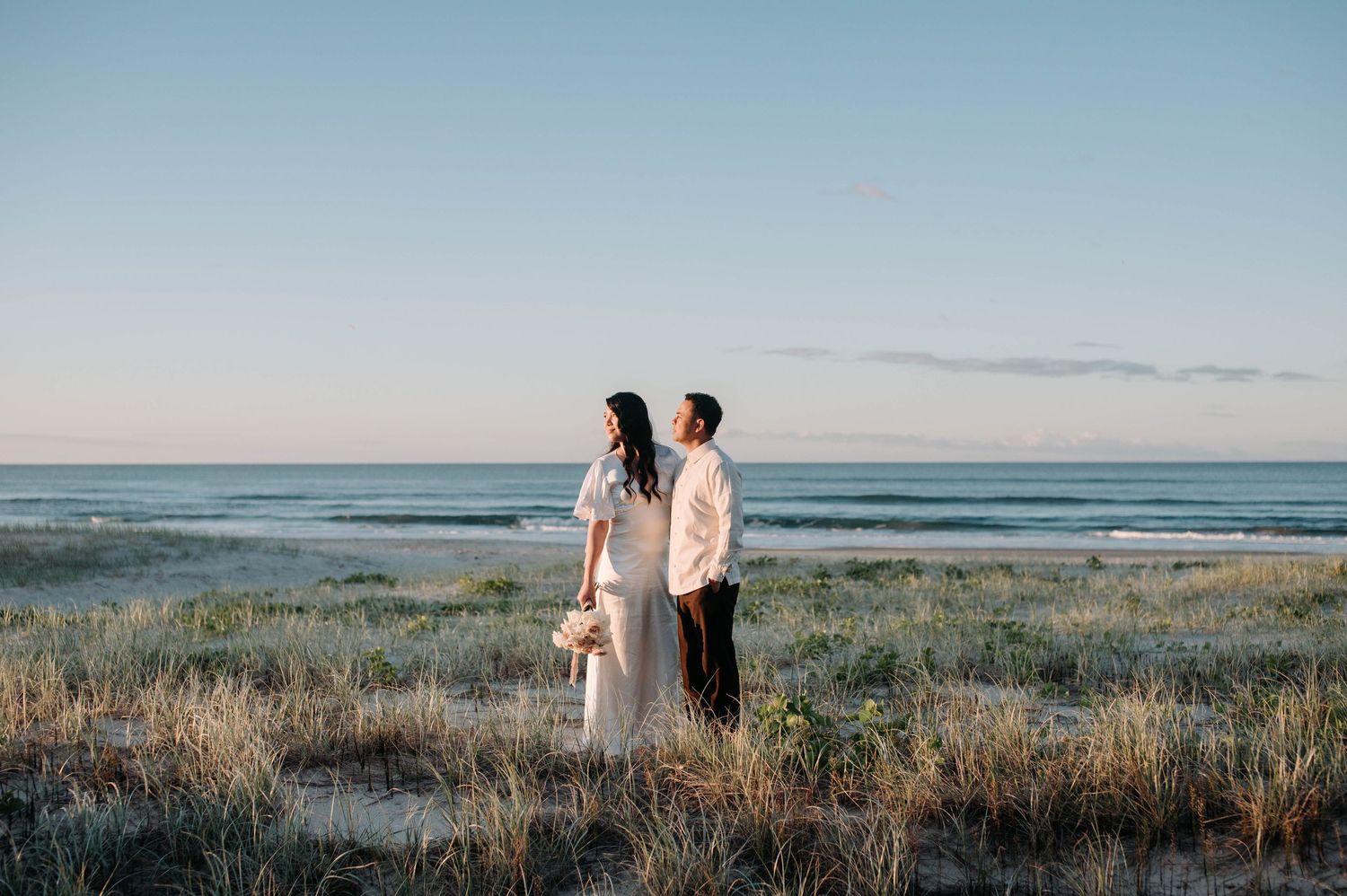 Two people stand together in flowing clothing against a scenic coastal landscape during golden hour.