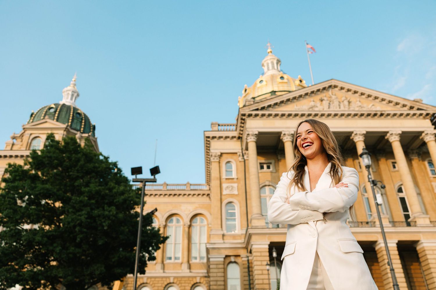 A young woman in a white pantsuit laughs, arms crossed confidently, in front of the Iowa State capitol.