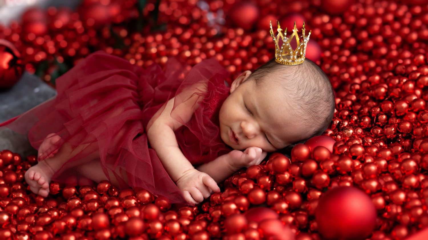 Sleeping newborn in burgundy dress wearing tiny crown lies among red Christmas ornaments and decorations.