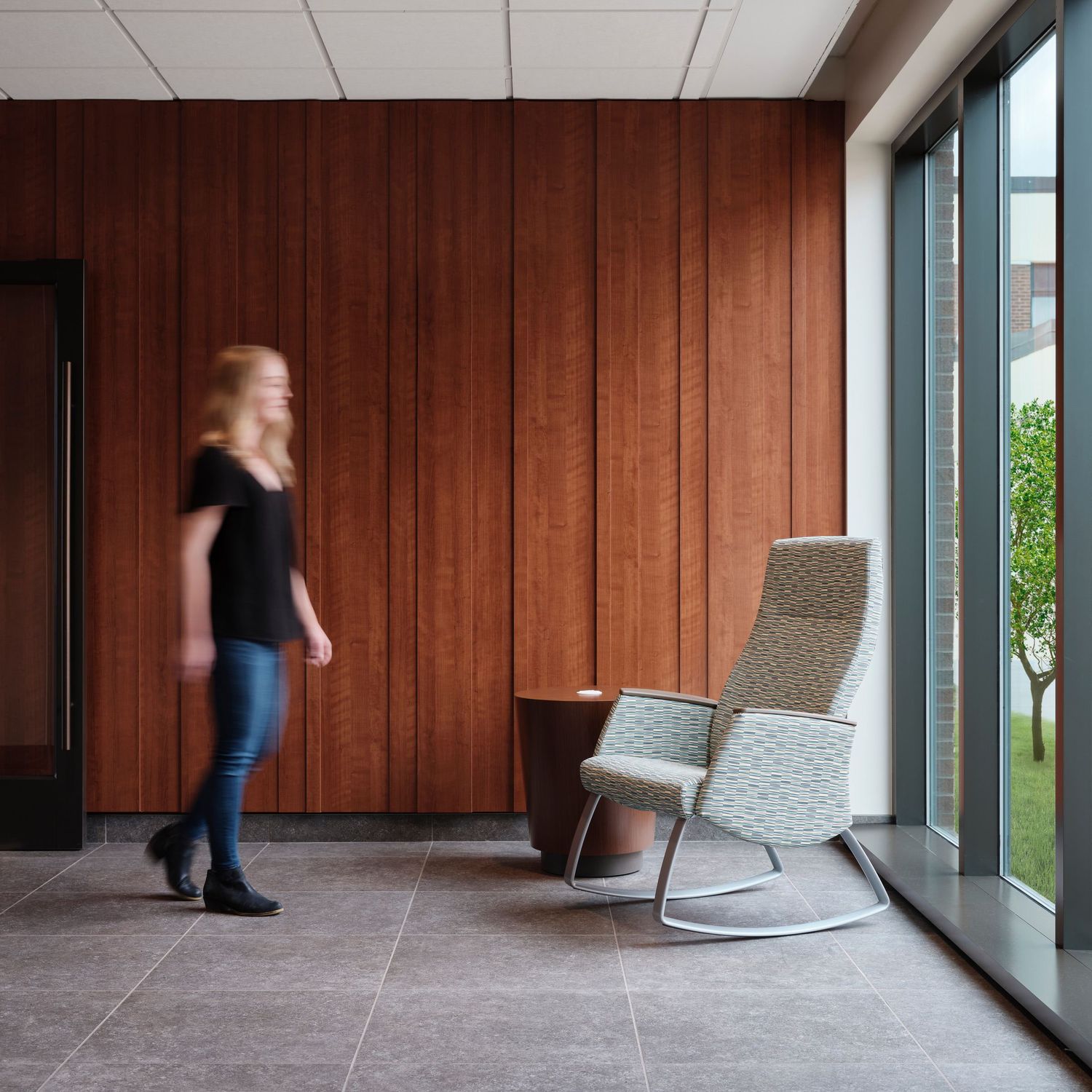 Modern waiting area with wood paneled walls, rocking chair, and large windows overlooking green space.