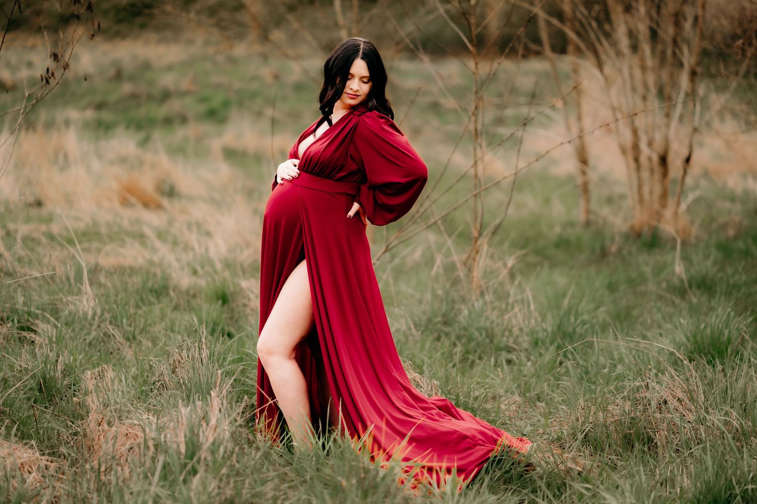 A flowing red velvet gown with long sleeves and side slit photographed in a rustic outdoor field setting for maternity photos in Tacoma.