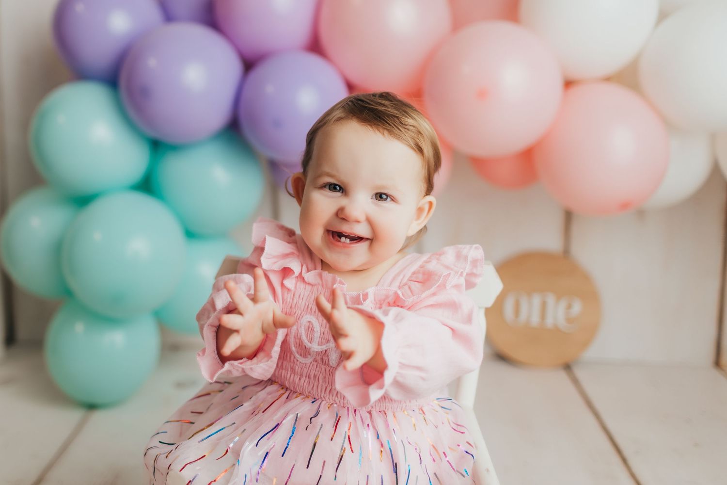 Happy baby wearing ruffled pink dress poses with colorful balloon display.