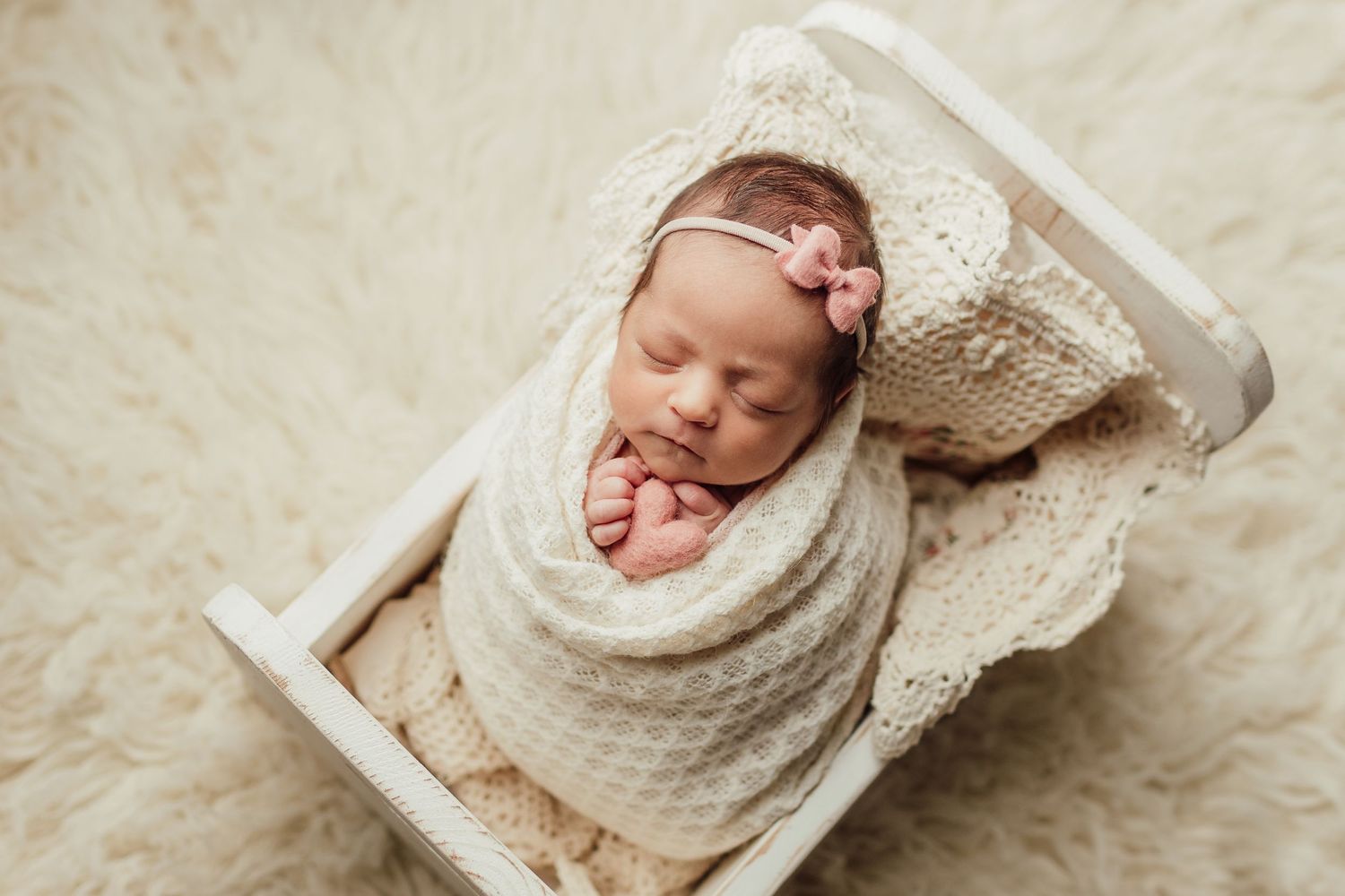 Sleeping newborn wrapped in cream knit blanket in tiny white bed.