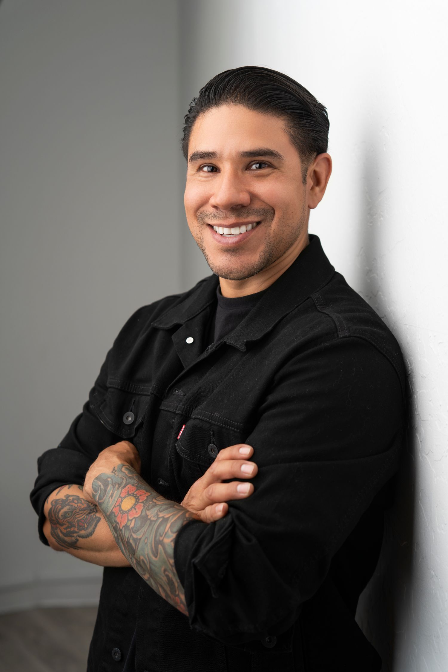 Professional portrait of person in black denim jacket with tattoo sleeve against gray background.