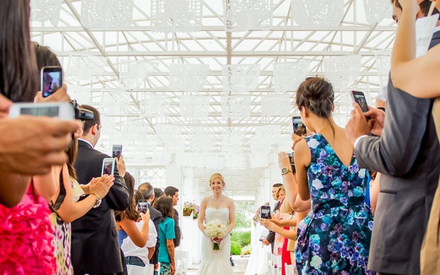 Wedding guests line aisle with phones capturing bride walking through industrial venue space.
