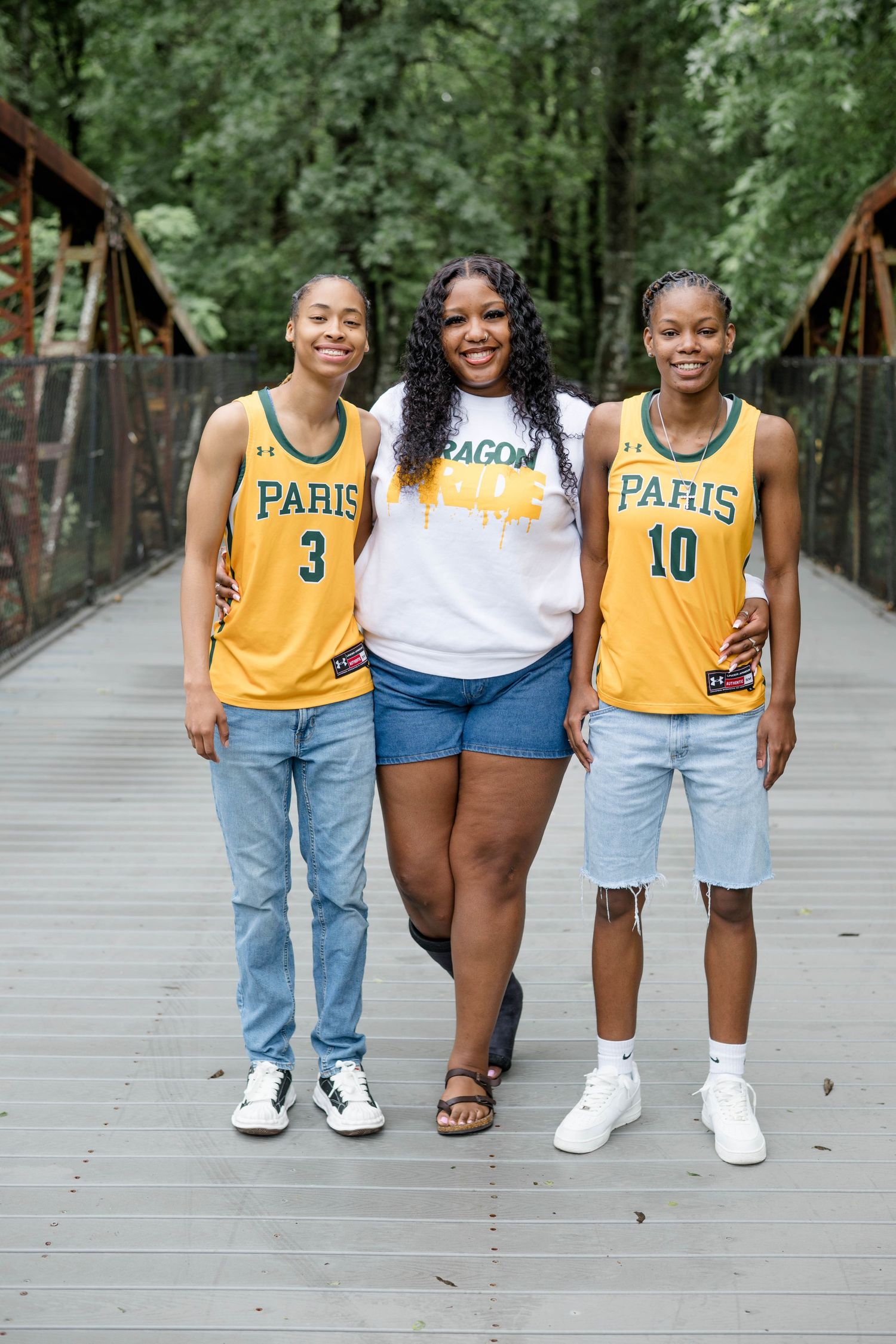 Three people in casual clothing pose together on a wooden bridge surrounded by green trees.