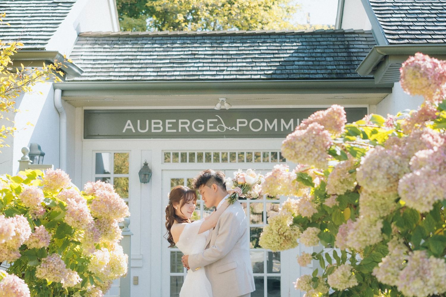 A couple embraces under the Auberge Pomme sign surrounded by blooming hydrangeas.