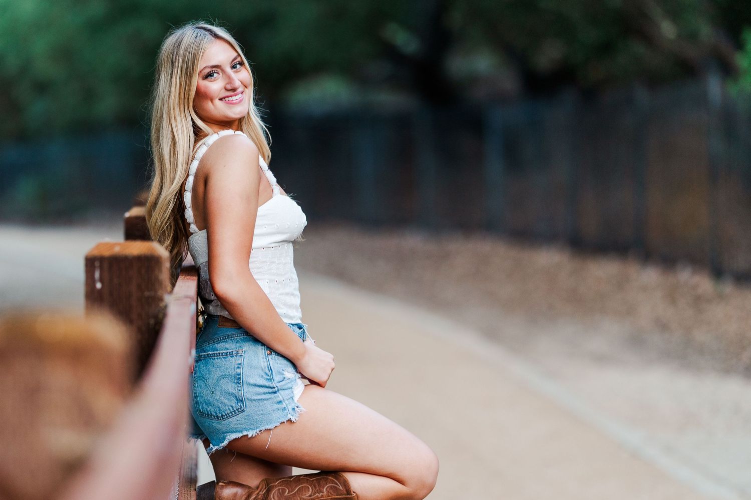 A person in a white tank top and denim shorts sits on a wooden railing along a walking path on a sunny day at Conejo Valley Botanic Garden.