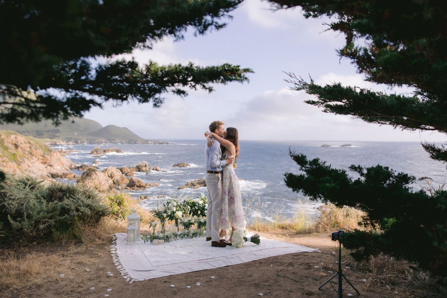 A couple shares a romantic moment on a wooden platform overlooking the Pacific Coast's rocky shoreline and crashing waves.