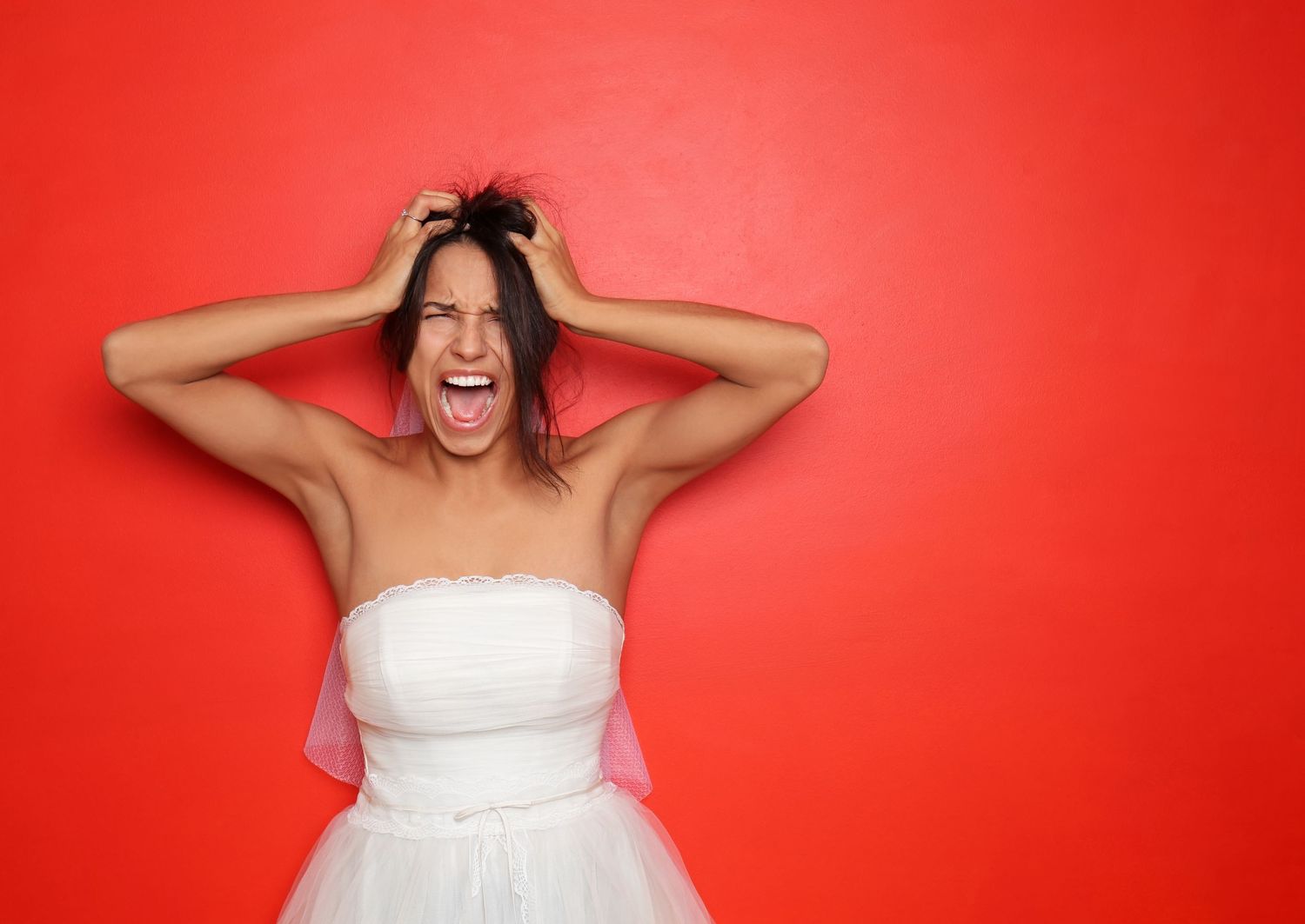 Person in white strapless dress putting hands on head and screaming against bright red background.