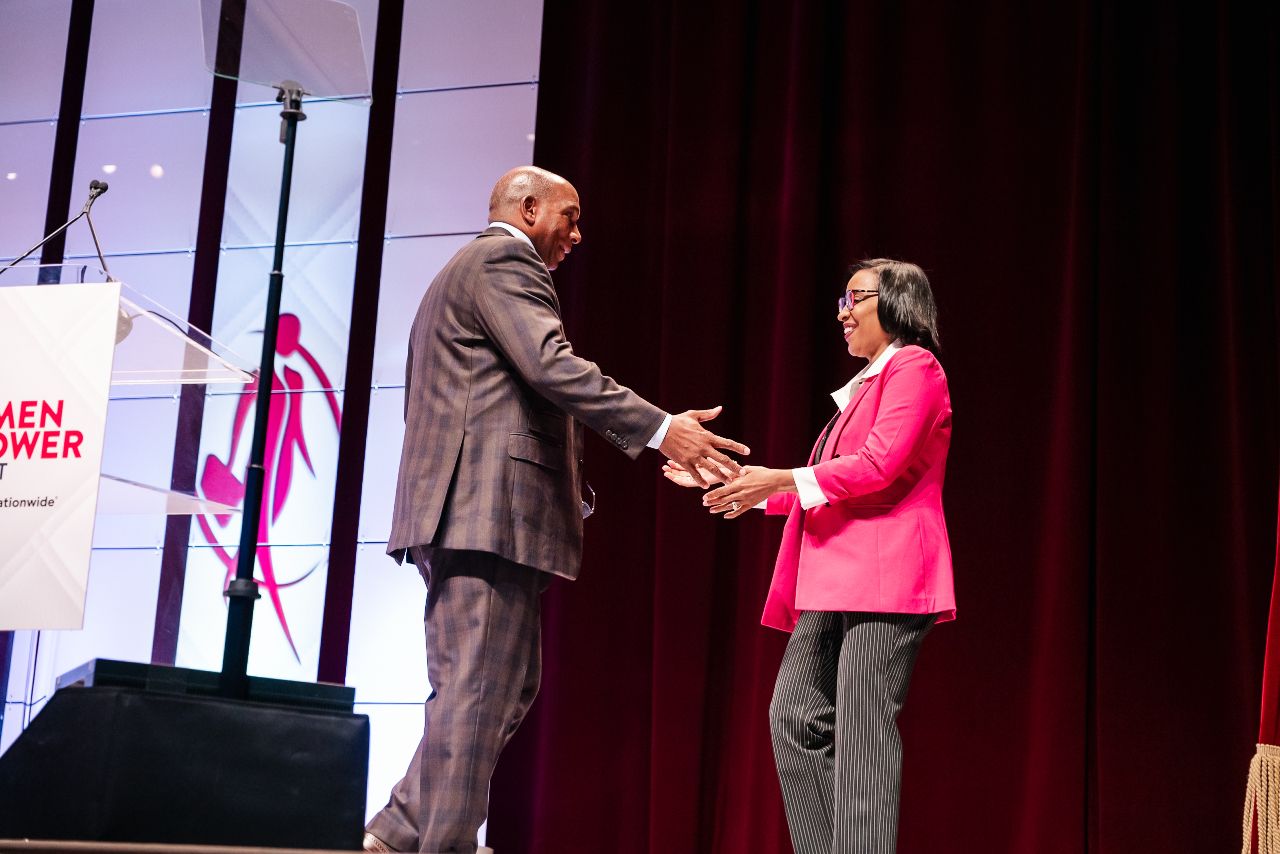 Two people shake hands on stage at a professional event with red curtains and spotlights in the background.