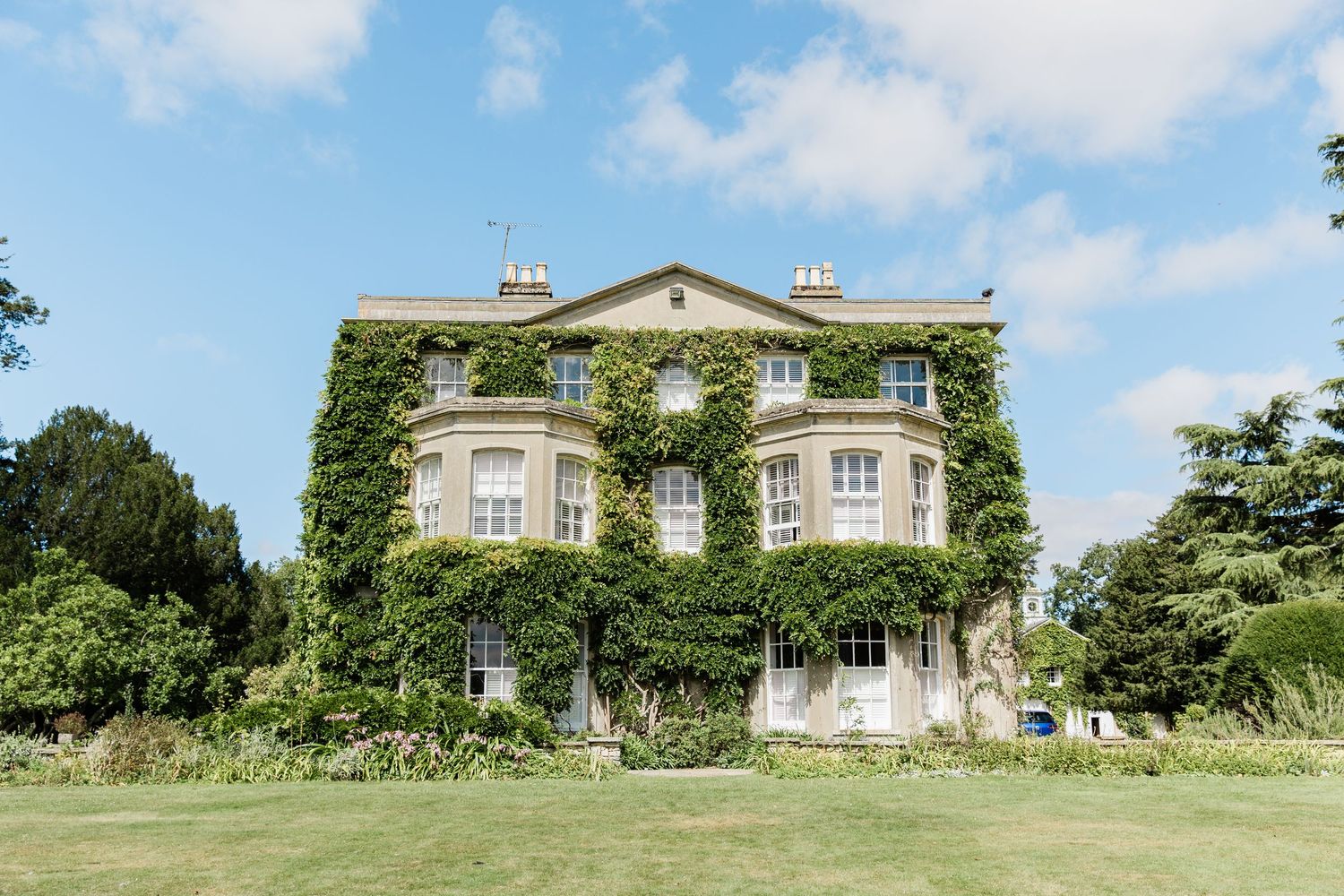 Elegant Georgian mansion covered in lush green ivy with bay windows and manicured lawn on a sunny summer day.