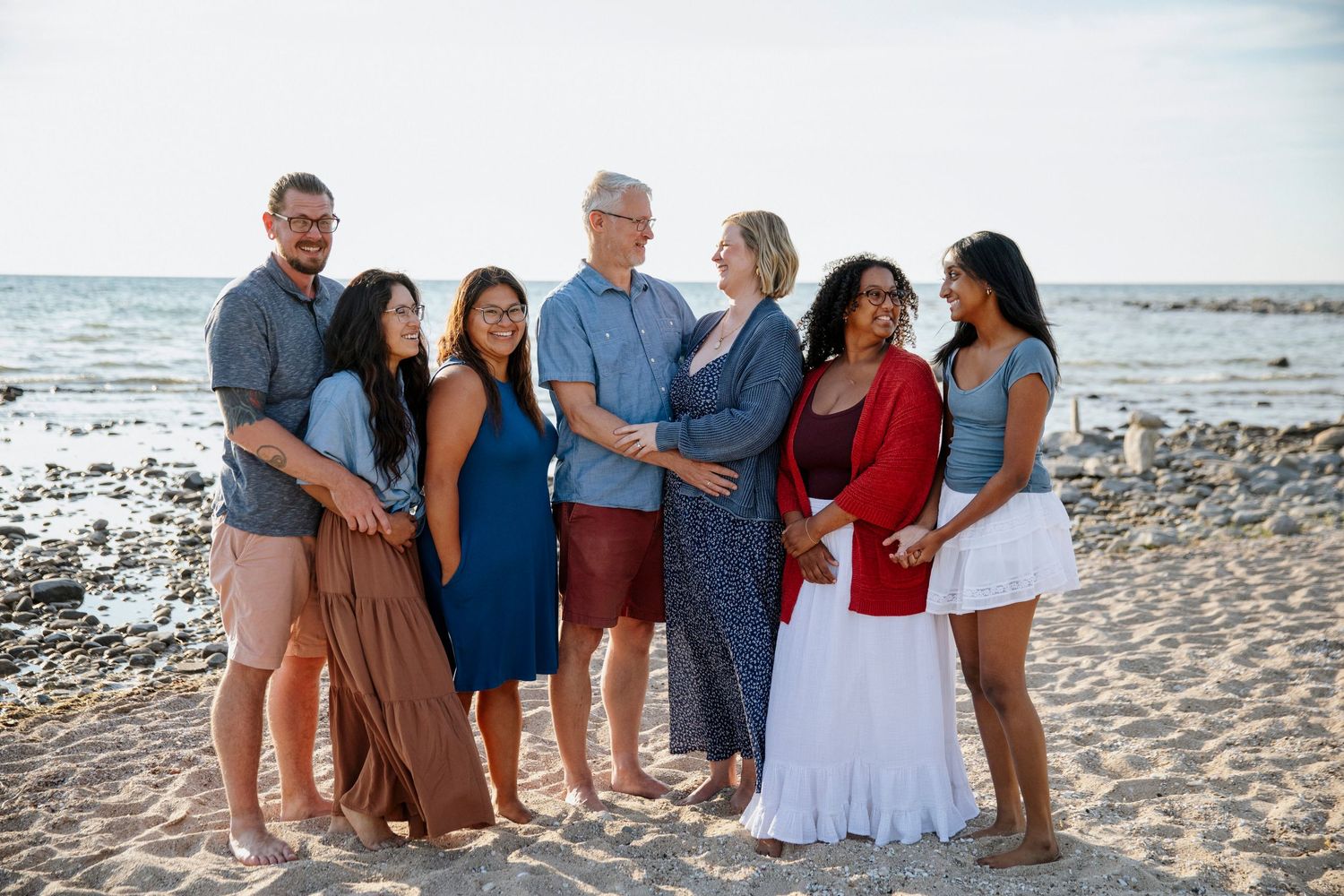 A joyful multigenerational family of seven poses together on a sandy beach with ocean waves in the background.