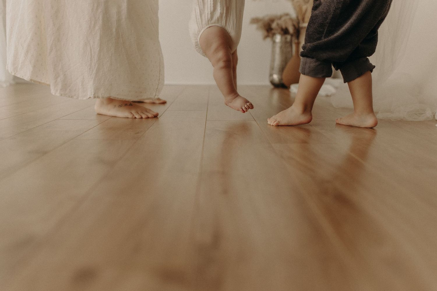 Barefoot adult and child standing on warm hardwood floor, captured from low angle in bright minimalist room.