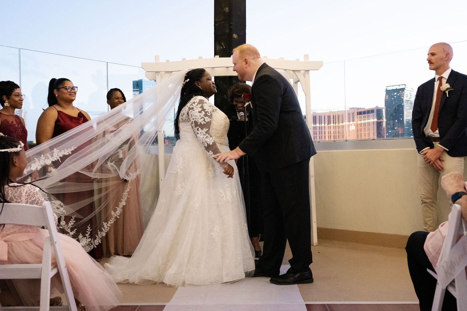 Bride in lace gown and veil exchanges vows with groom at outdoor rooftop wedding ceremony with city skyline backdrop.