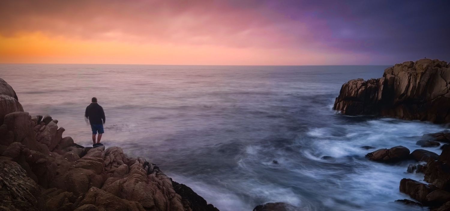 Lone figure stands on rocky coastal cliffs at sunset, gazing over dramatic crashing, long-exposure waves under a vivid purple & orange sky.
