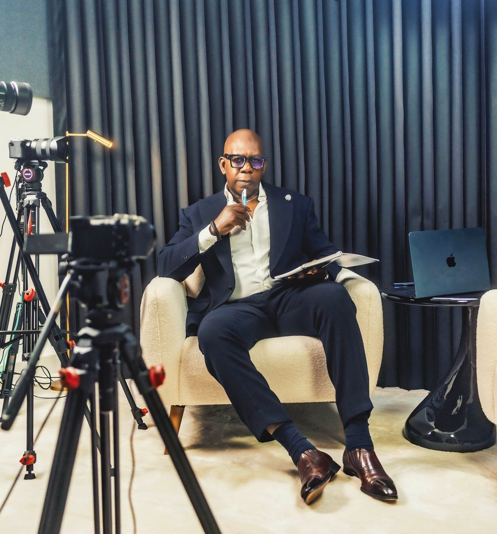 Professional man in suit sitting in armchair during video recording session with camera equipment and laptop.