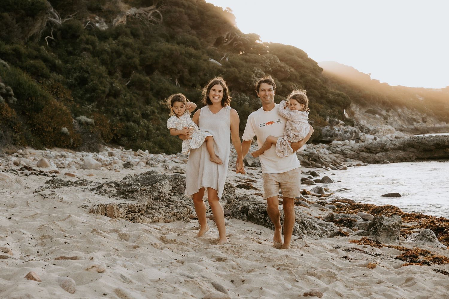 Family of four walking barefoot on sandy beach, parents carrying toddlers in white outfits, rocky cliffs in background.