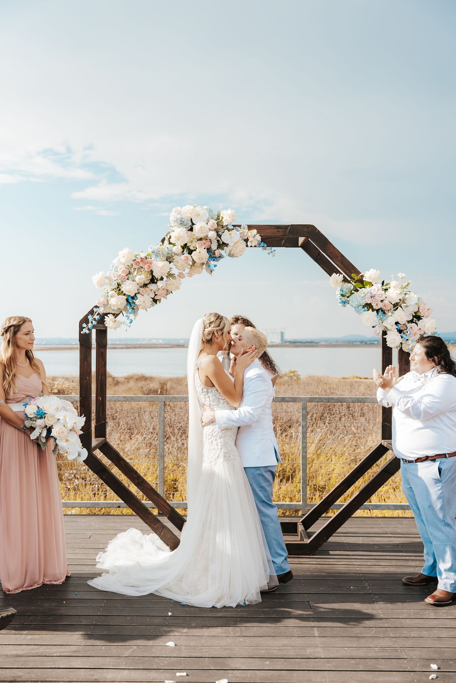 Bride and groom share first kiss under floral hexagonal arch on beachside deck with bridesmaid and officiant nearby.