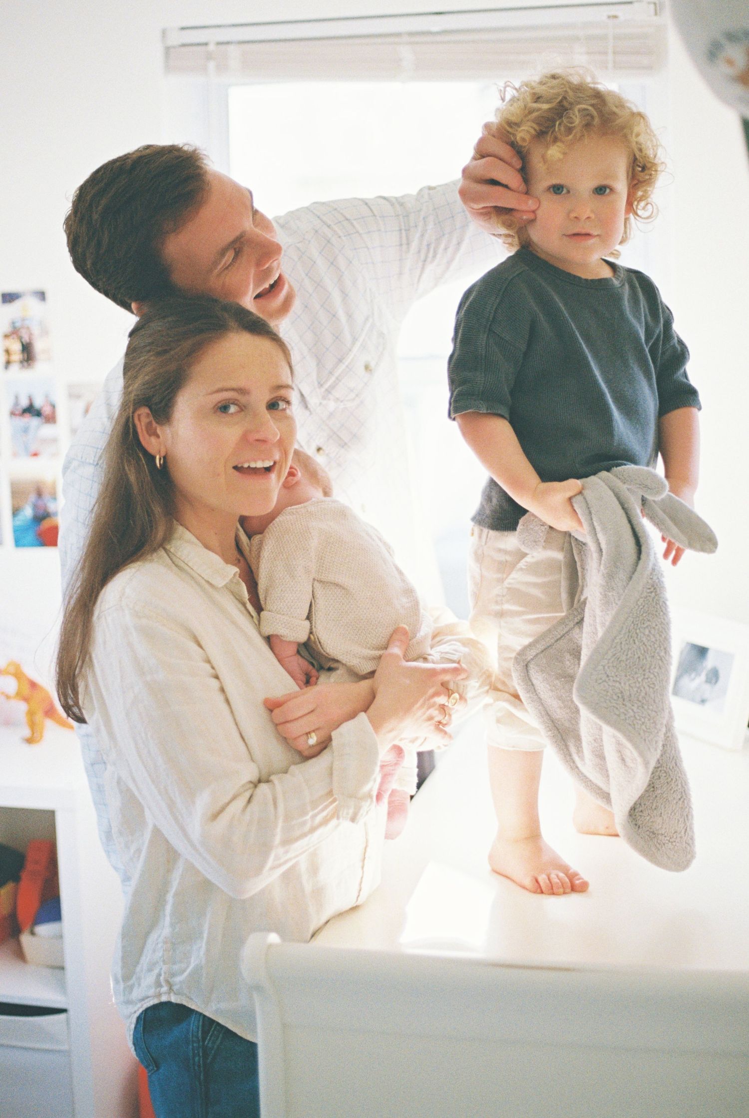 Happy family with curly-haired toddler meeting newborn baby, parents smiling, colorful blocks on wooden table near window.