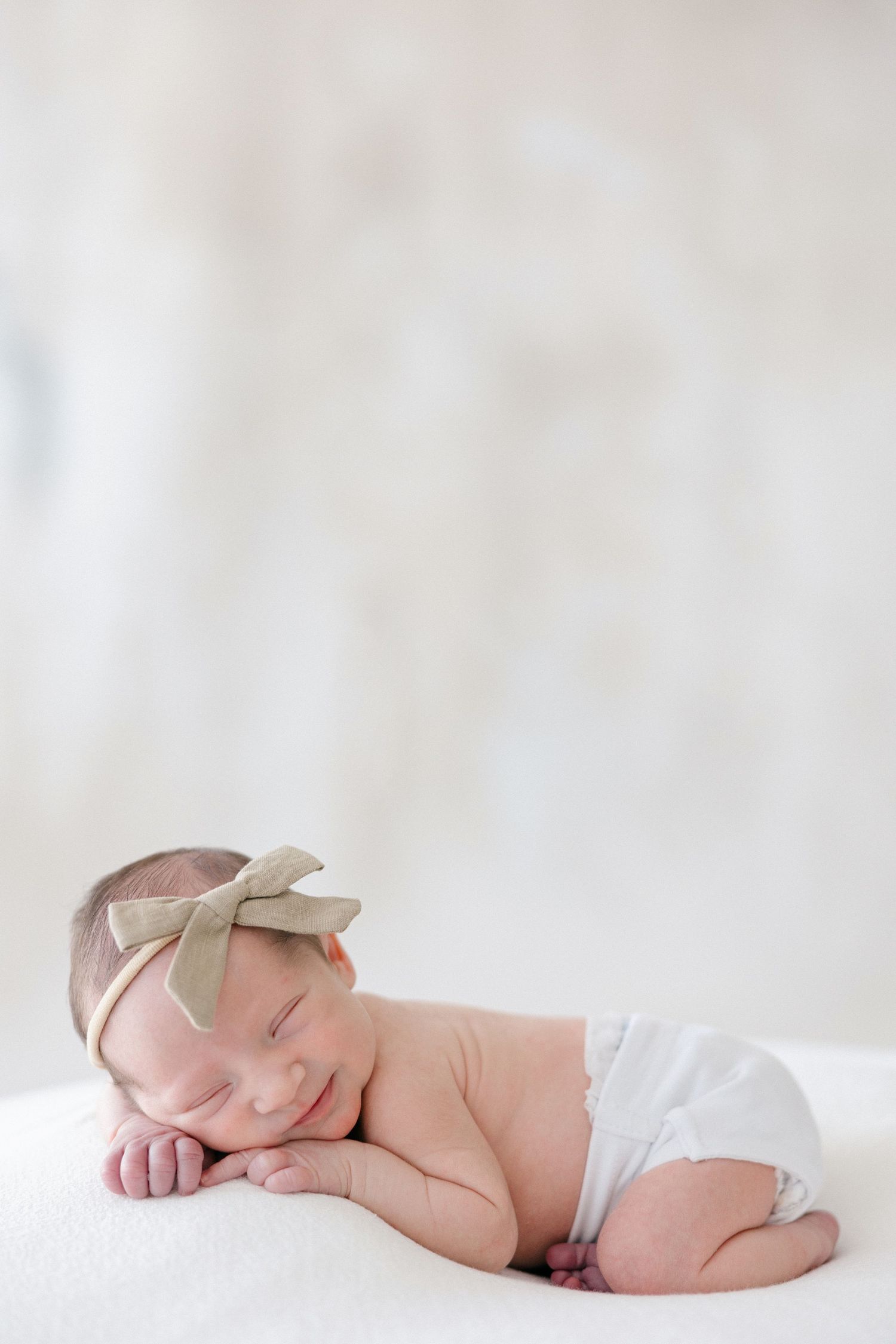 Sleeping newborn baby wearing white diaper and beige bow headband rests peacefully on soft white blanket.