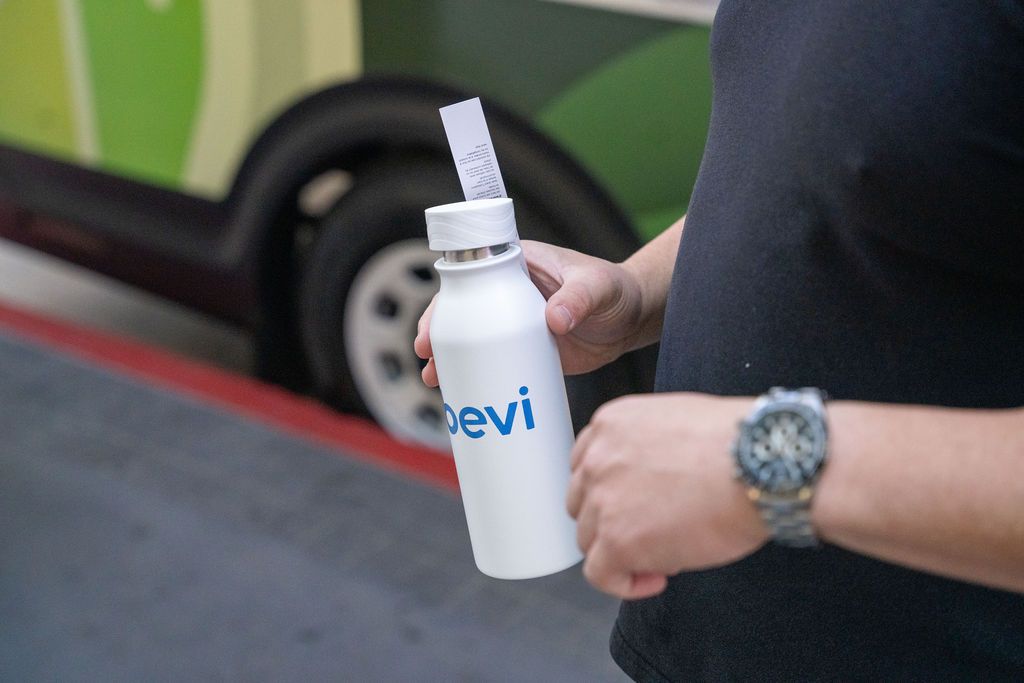 Person holding a white Bevi branded reusable water bottle outdoors near a green vehicle.