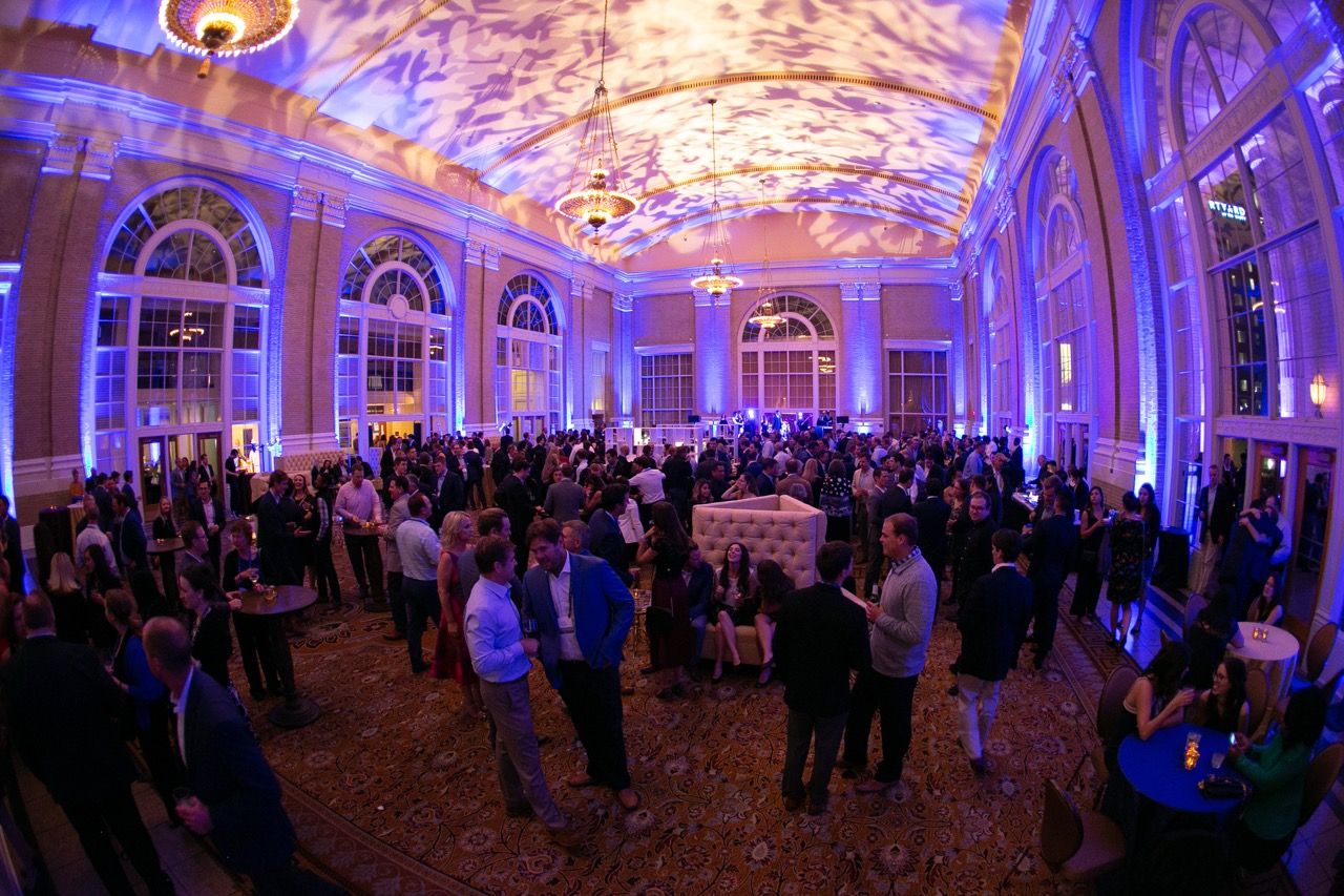 Elegant ballroom event with crowds of guests mingling under ornate ceilings with blue and purple ambient lighting.