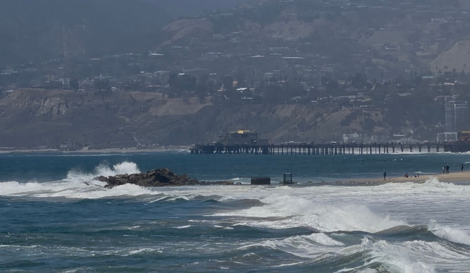 Rough ocean waves crash near a pier with misty hills in the background on a cloudy coastal day.