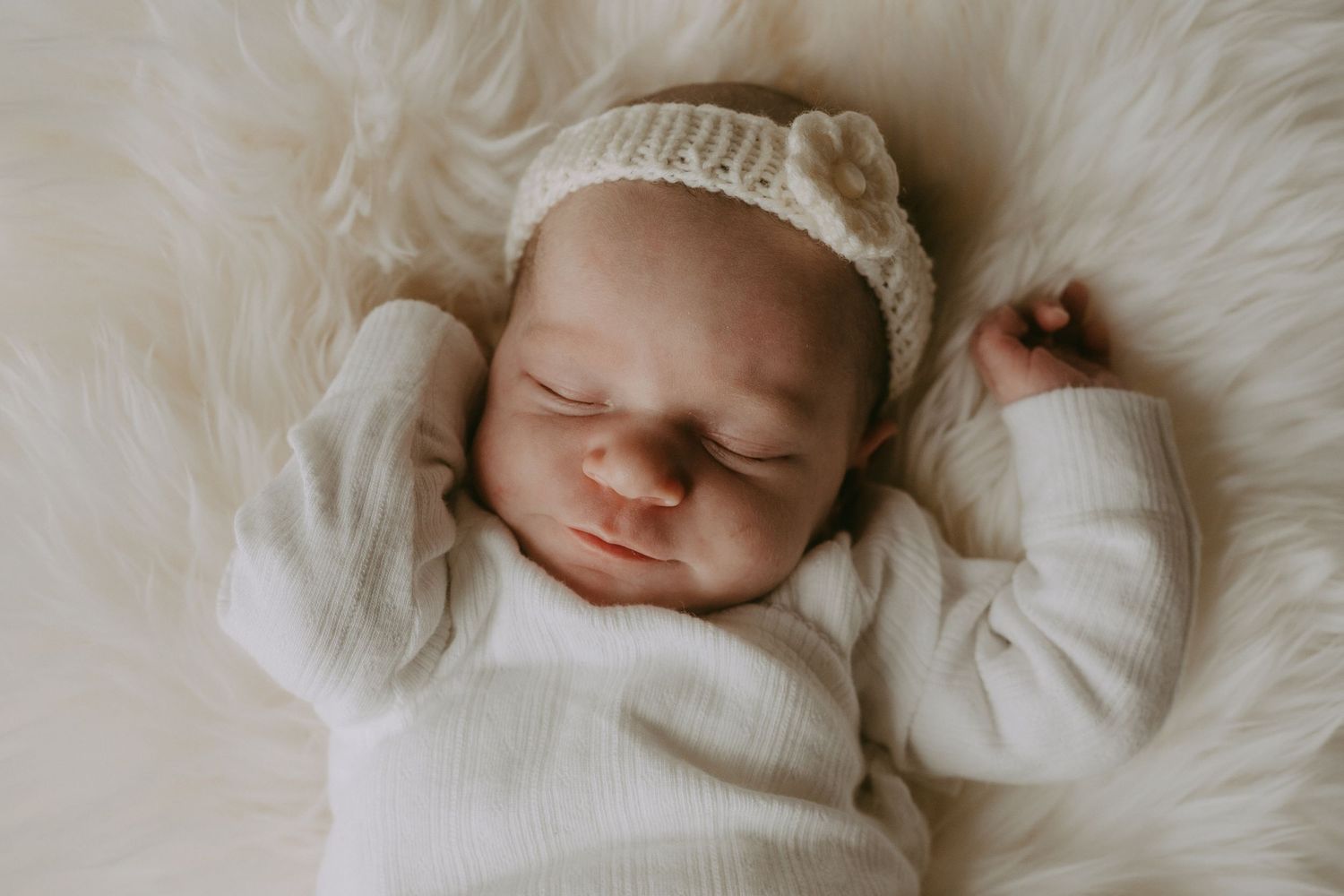 Peaceful newborn baby sleeping on fluffy white fur, wearing a cream knit outfit and matching bear ear hat.
