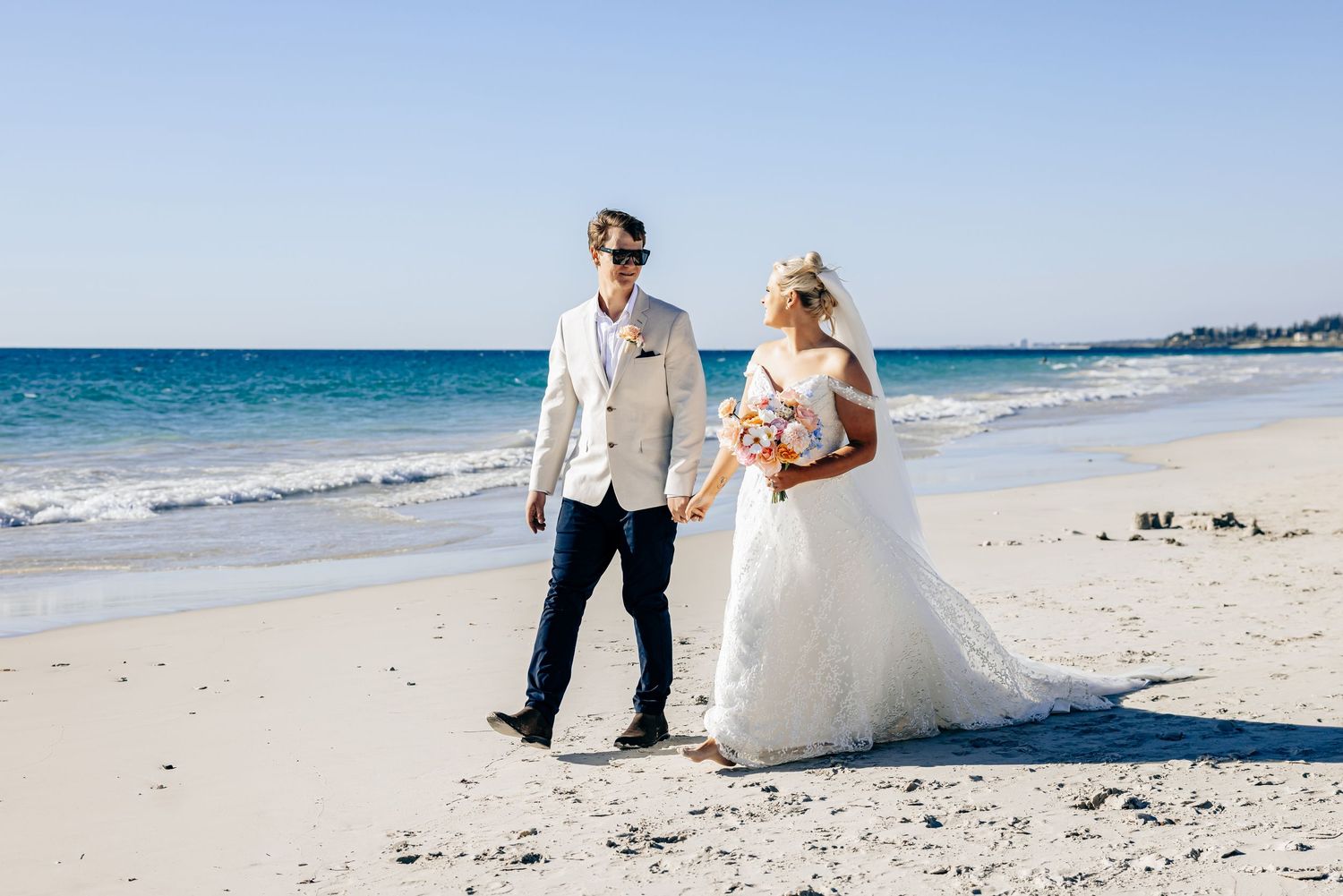 Bride and groom walking hand in hand on a sunny beach, bride in white gown holding bouquet, groom in tan suit.