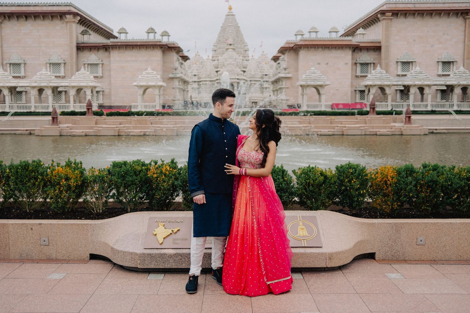 Couple in traditional Indian attire posing in front of an ornate white temple with reflecting pool.