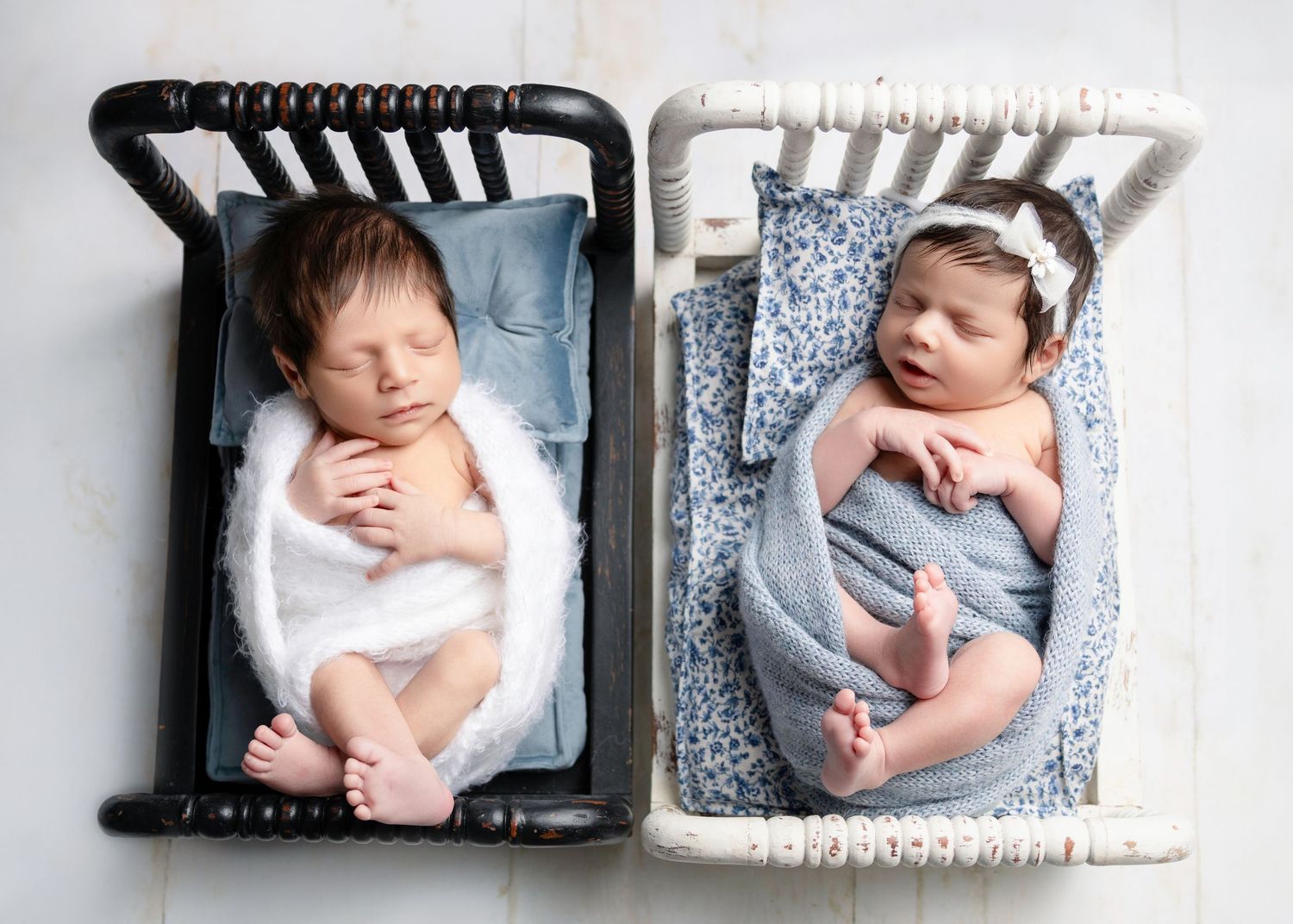 Two sleeping newborn twins in miniature cribs, one in black and one in white, wrapped in soft blue blankets.