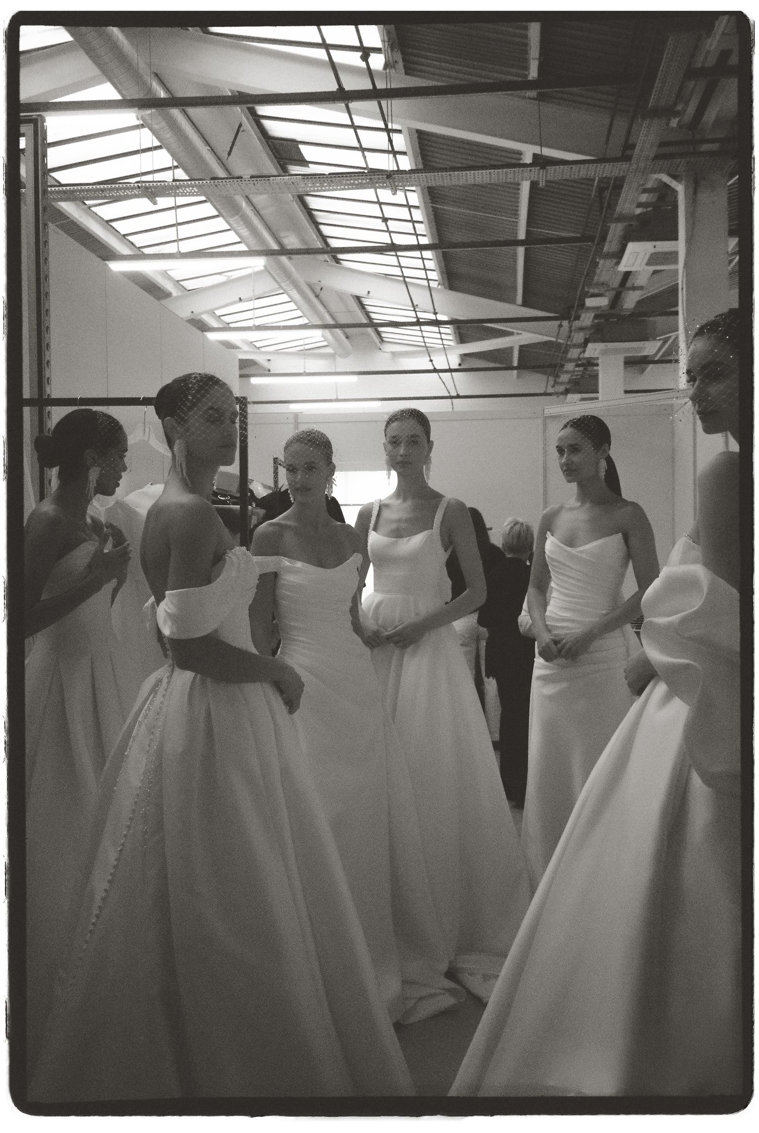 Black and white photo of models in elegant white bridal gowns gathered backstage at a wedding fashion show.