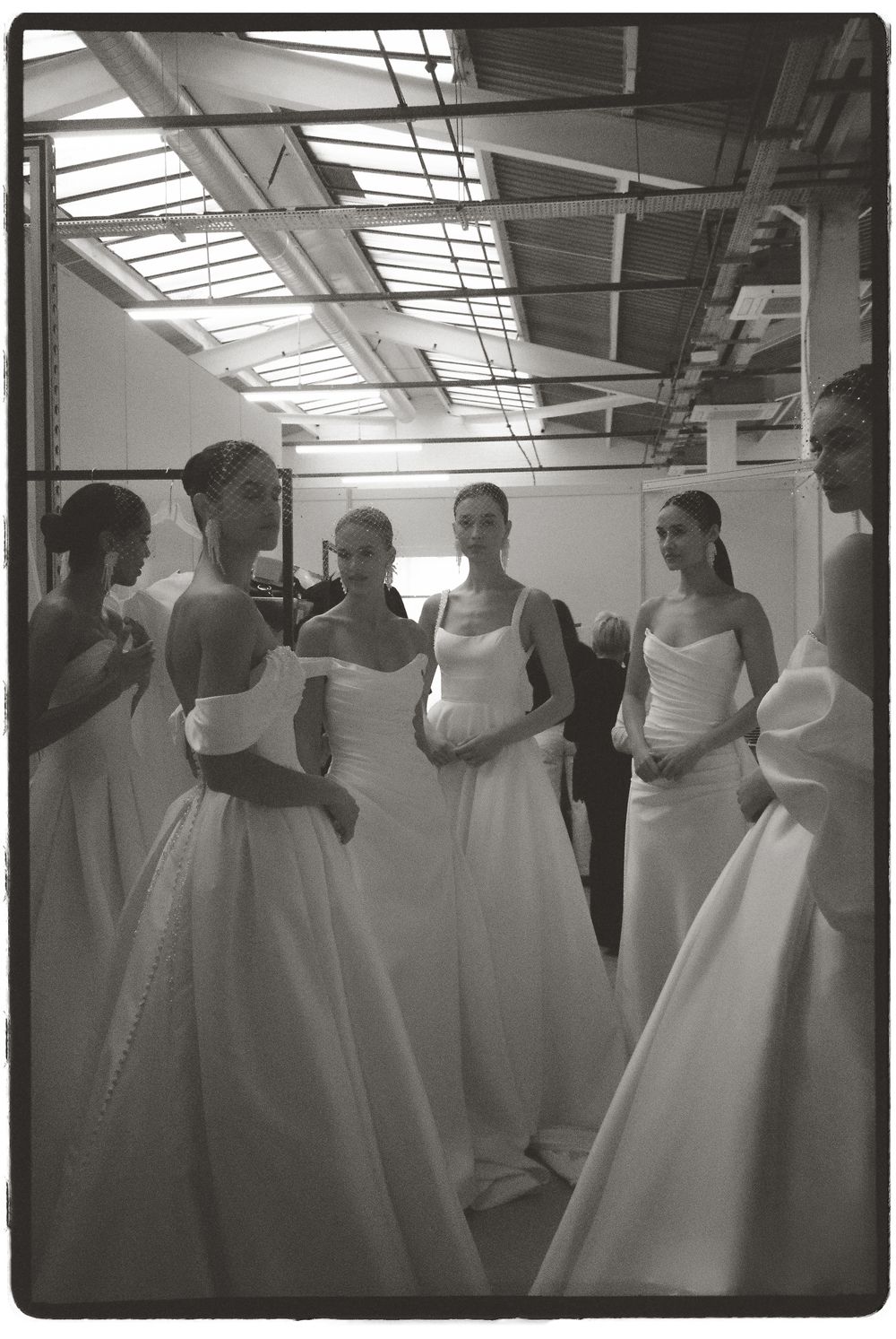 Black and white photo of models in elegant white bridal gowns gathered backstage at a wedding fashion show.