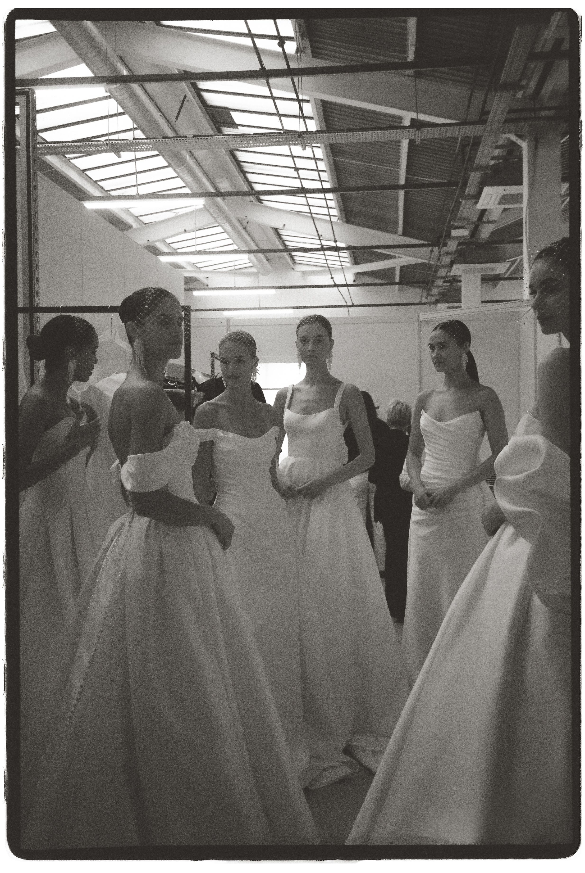 Black and white photo of models in elegant white bridal gowns gathered backstage at a wedding fashion show.