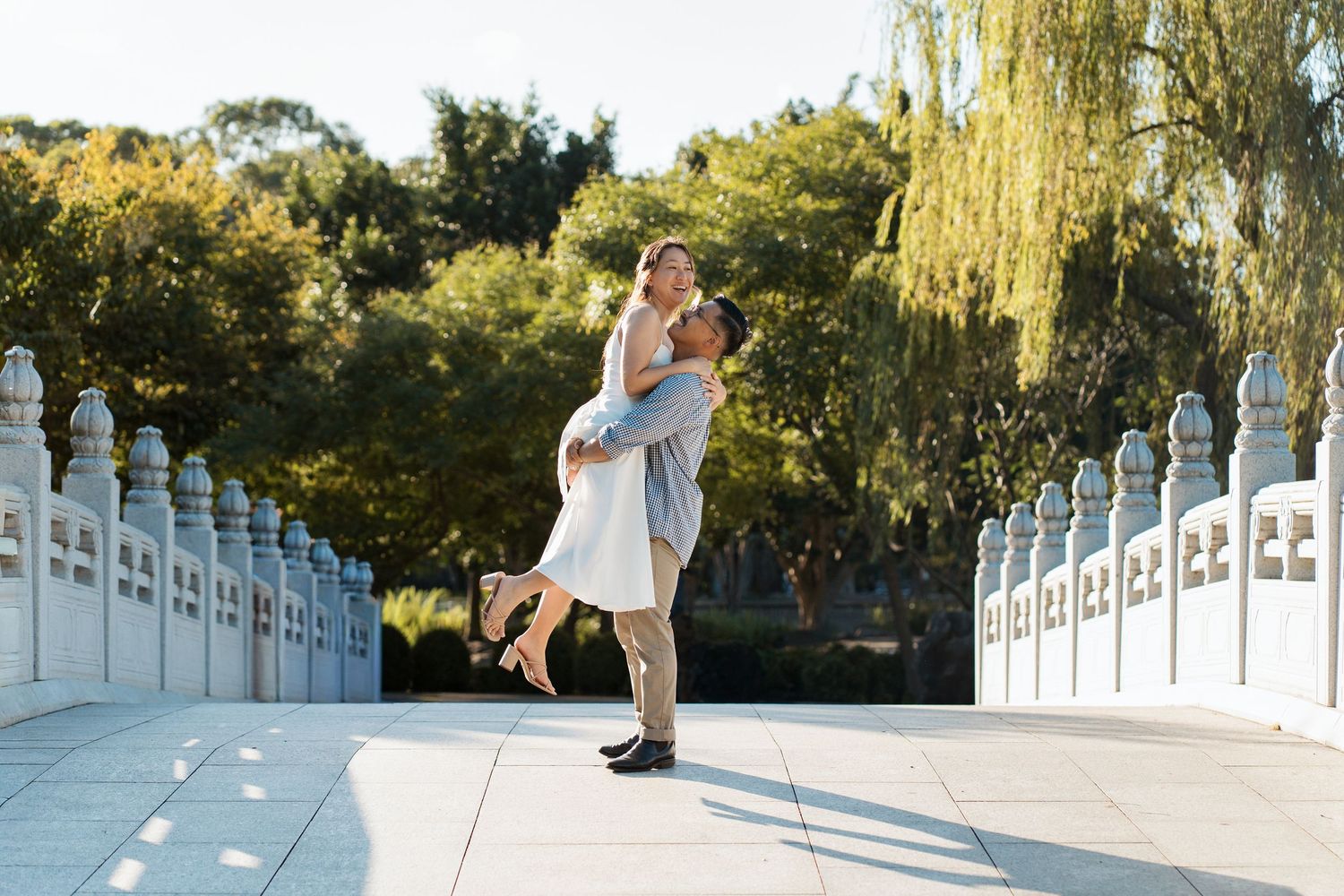Couple sharing a romantic moment on a white bridge, man lifting woman in a sunny Nurragingy park with green trees during couple photoshoot