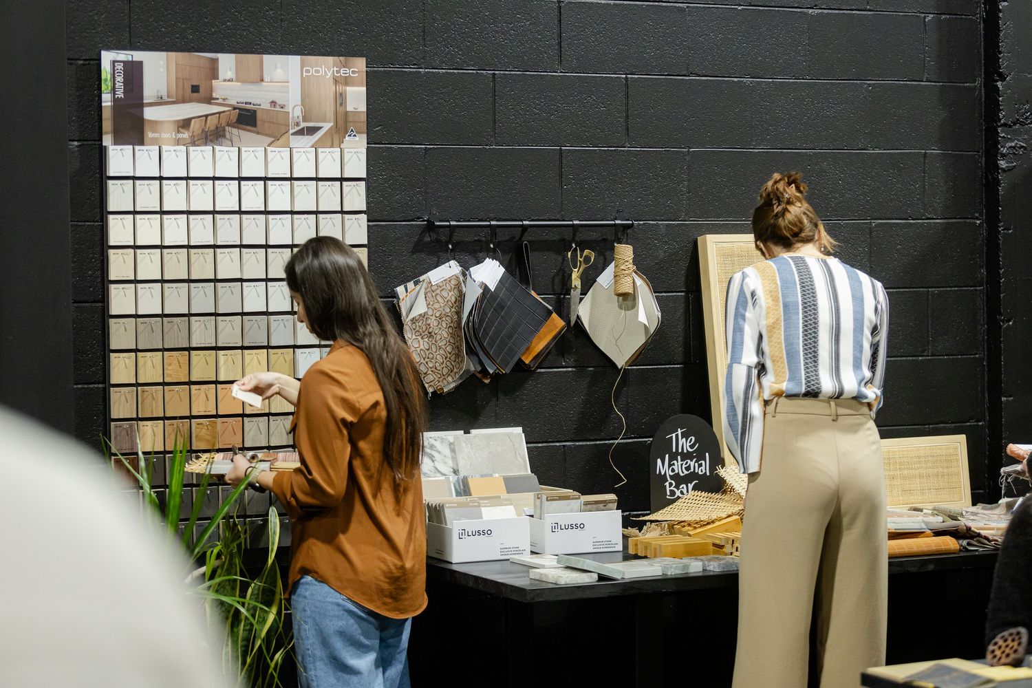 Two women browse material samples and decor displays at an interior design trade show booth.