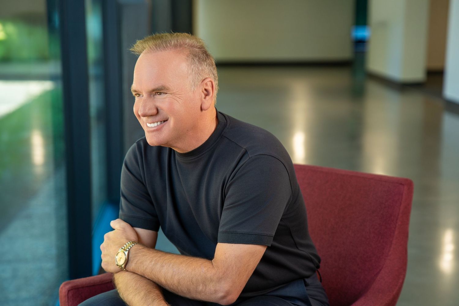 Smiling middle-aged man in dark t-shirt sitting relaxed in red chair, wearing gold watch, in modern bright interior.
