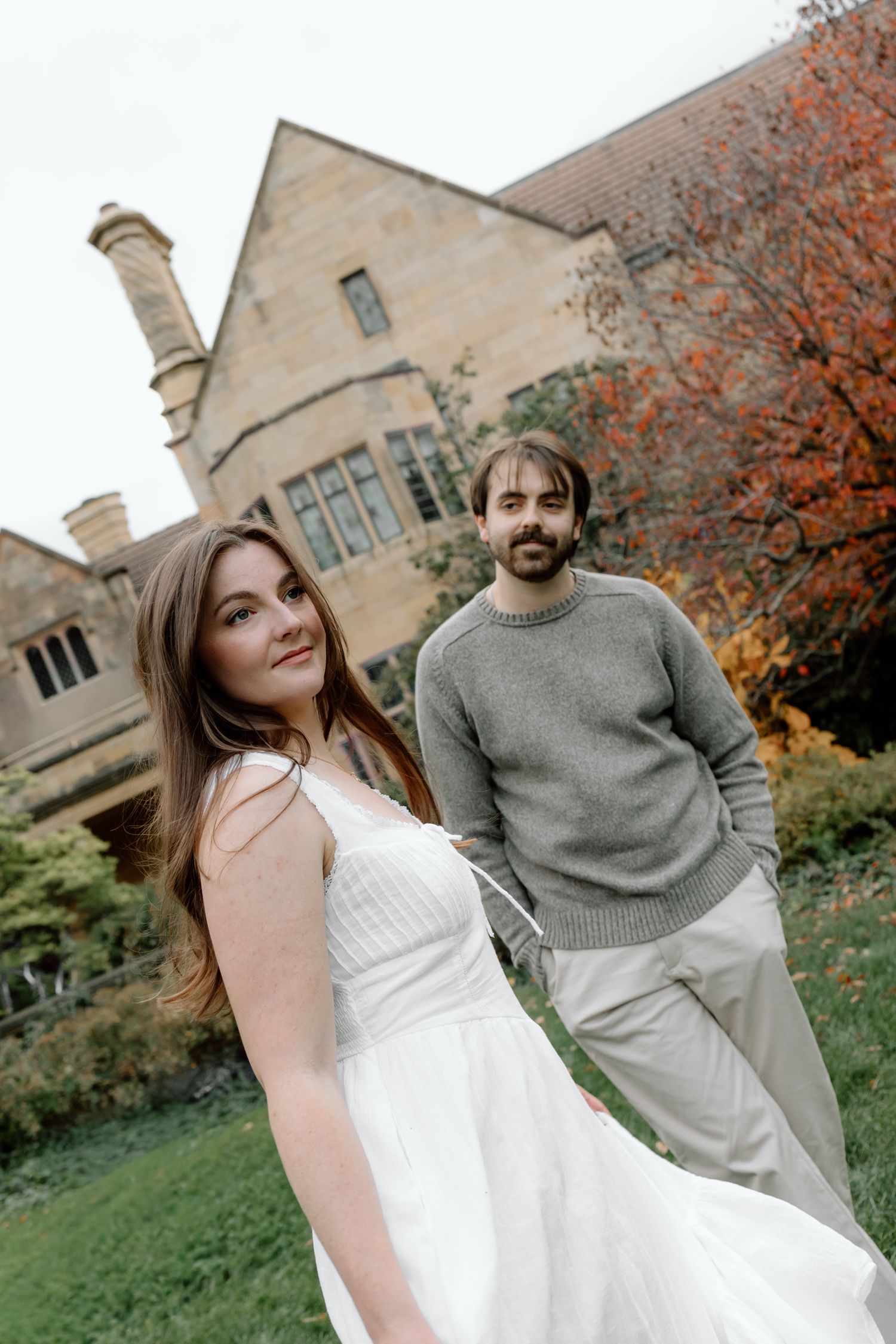 A couple poses in front of a stone manor surrounded by autumn foliage, woman in white dress, man in grey sweater.