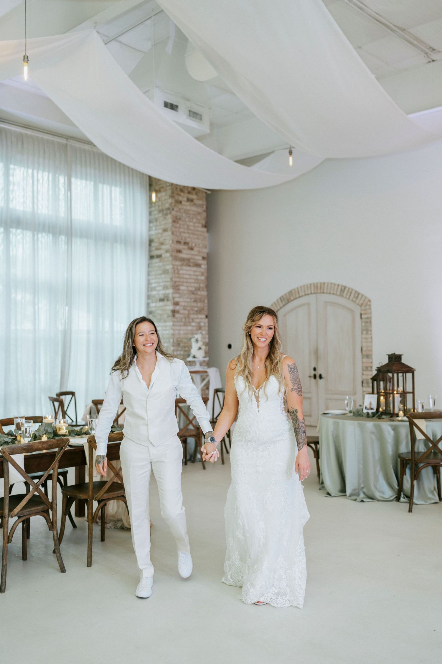 Two brides walk hand-in-hand at their wedding reception, one in a white suit and one in a lace gown.