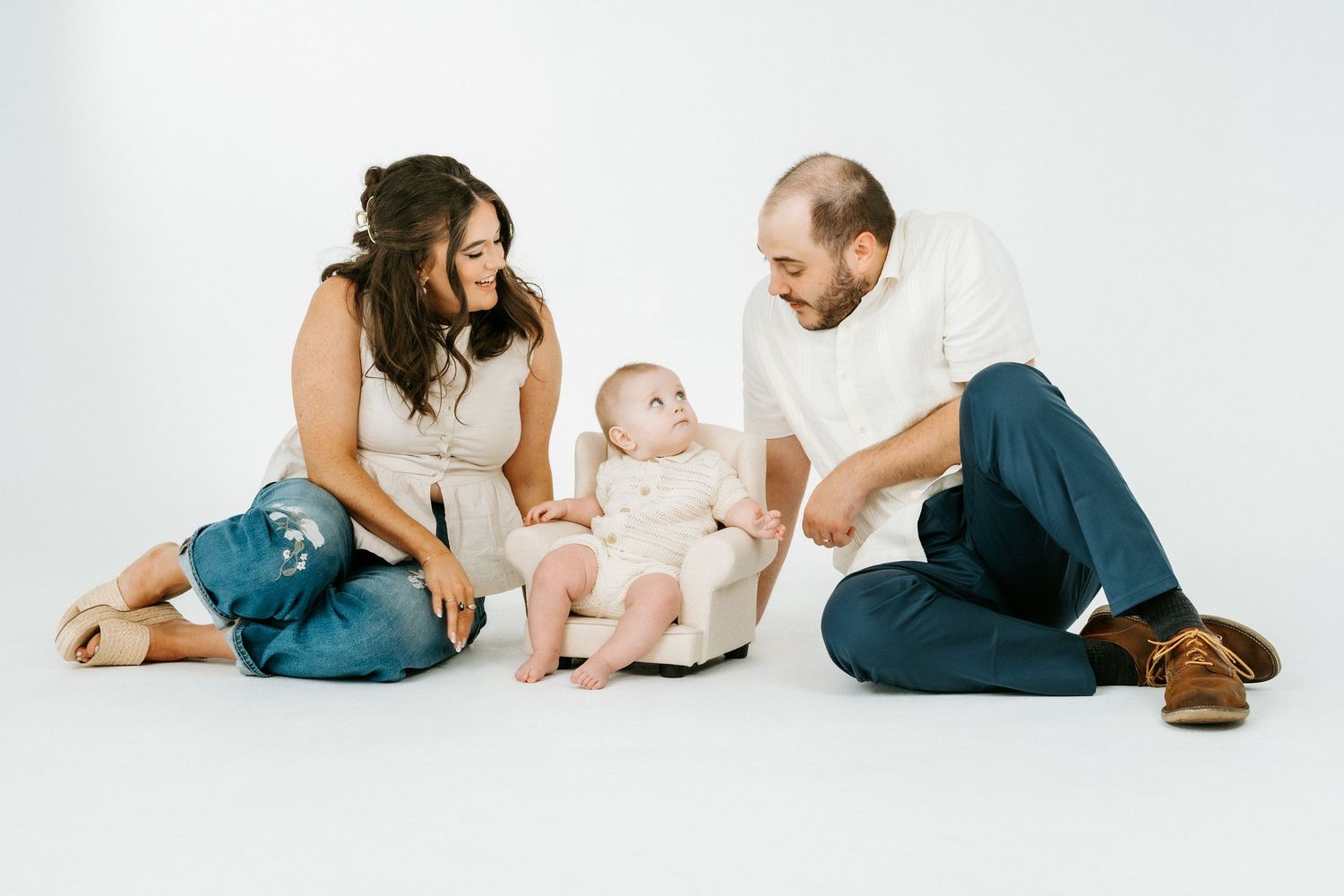 A young family with a baby sitting in a small chair between smiling parents on a white studio background.