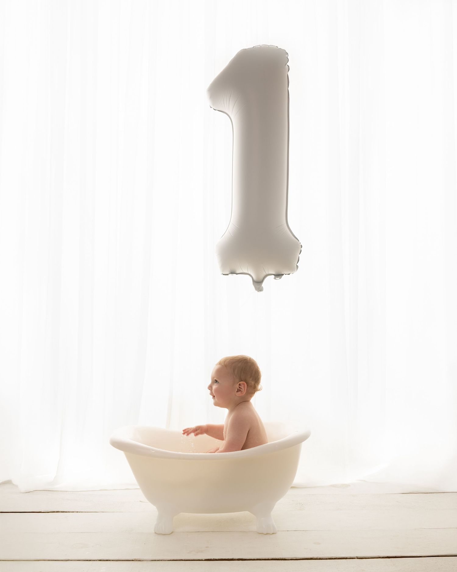 Baby sitting in a white clawfoot bathtub beneath a number one balloon, celebrating their first birthday.