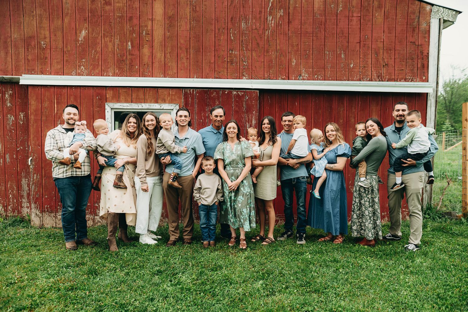 Large family group portrait standing in front of a rustic red barn on a green lawn.