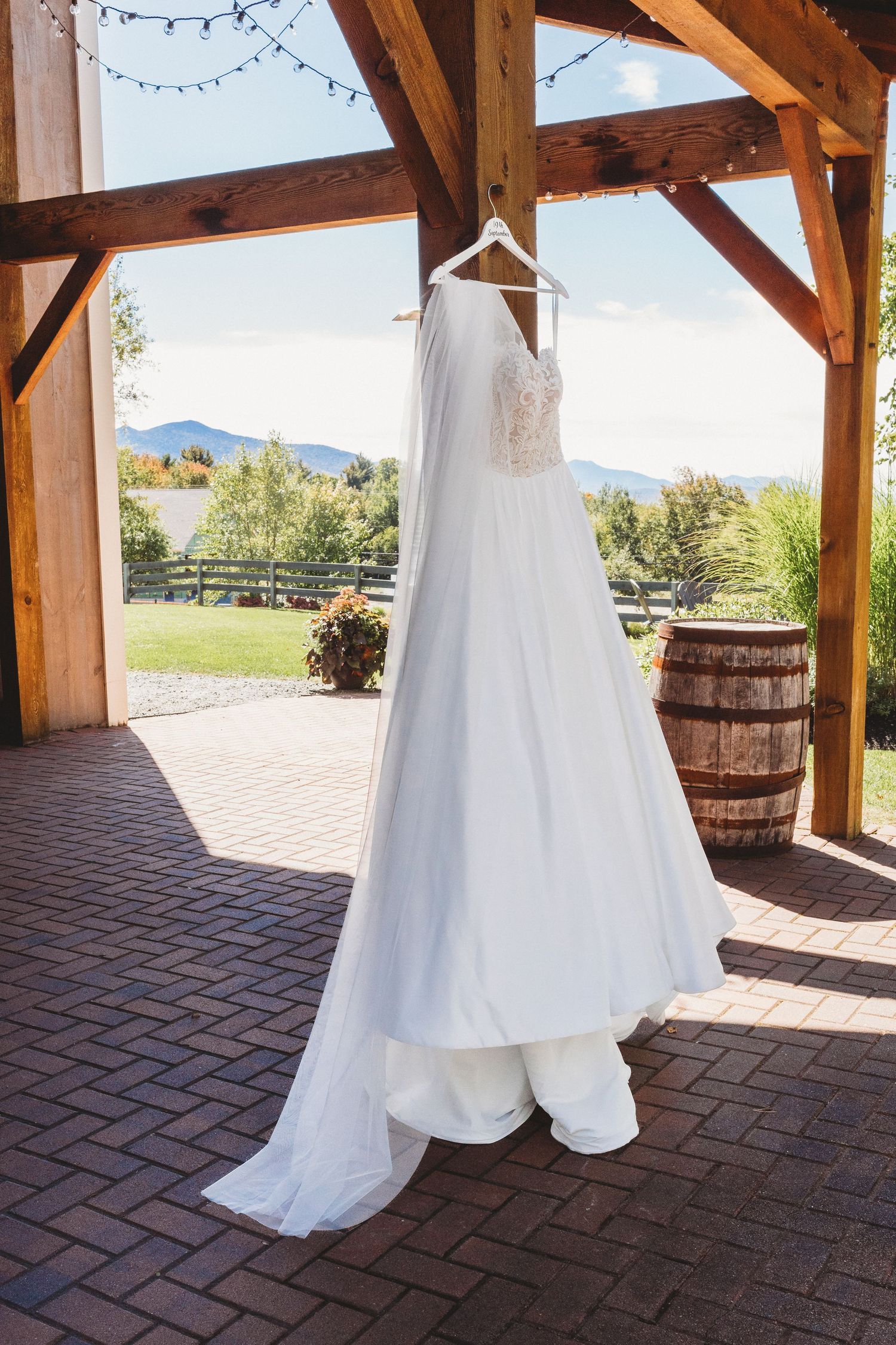 Wedding dress hanging from a wooden beam beneath a covered outdoor pavilion.