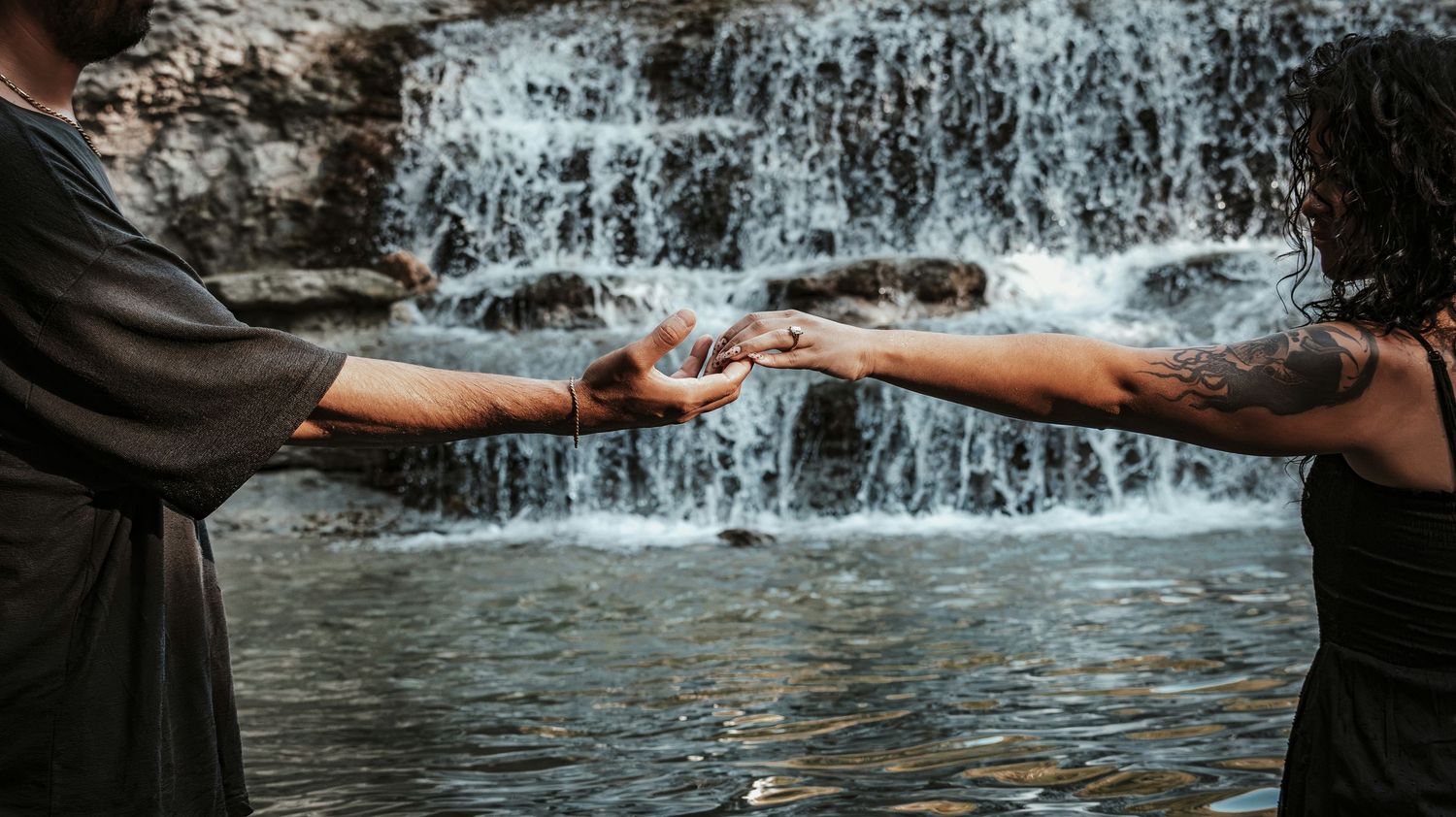 Two tattooed hands gently touching fingertips in front of a serene waterfall backdrop.