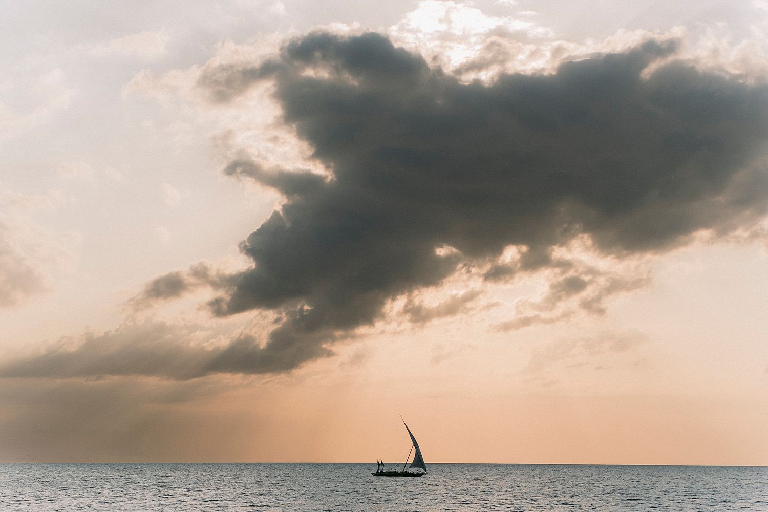 A lone sailboat glides across calm ocean waters beneath dramatic storm clouds at golden sunset.
