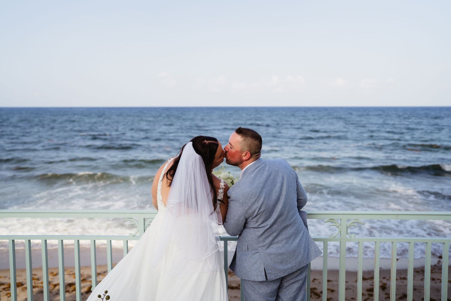 Bride and groom walk hand-in-hand along a lush garden path beside a white resort with palm trees and ocean views.